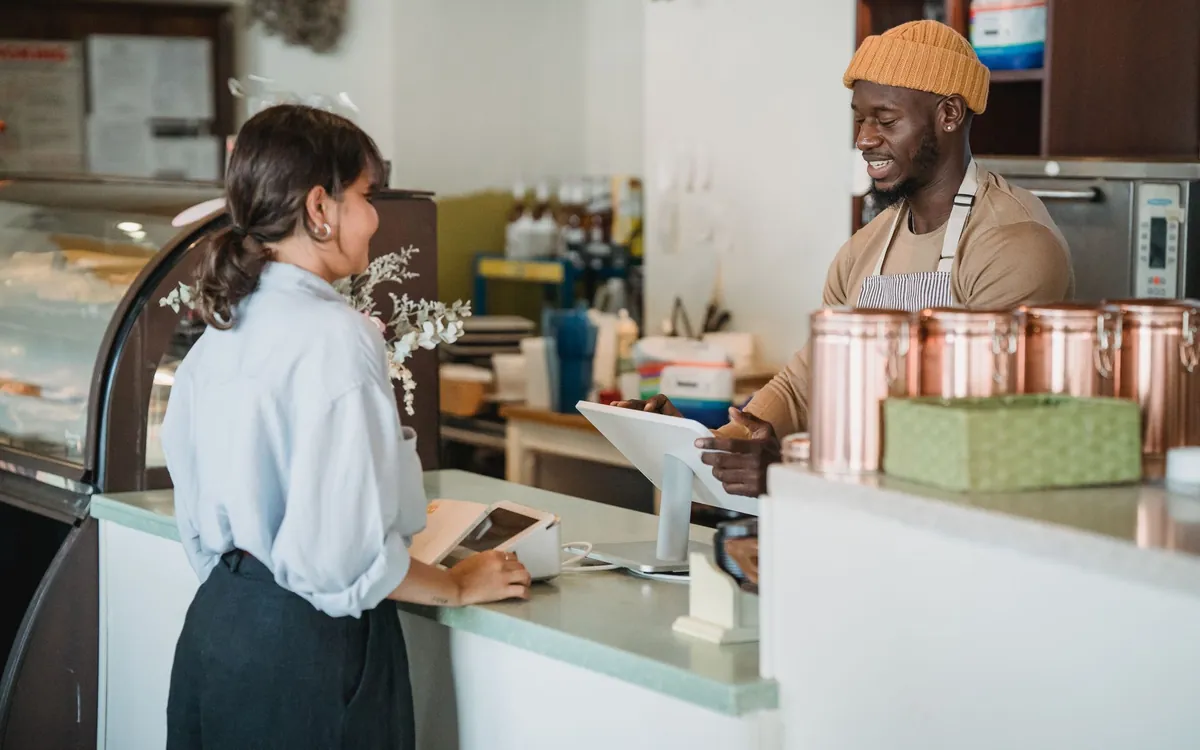 A customer paying at a cafe counter while a smiling barista rings up the order on a tablet